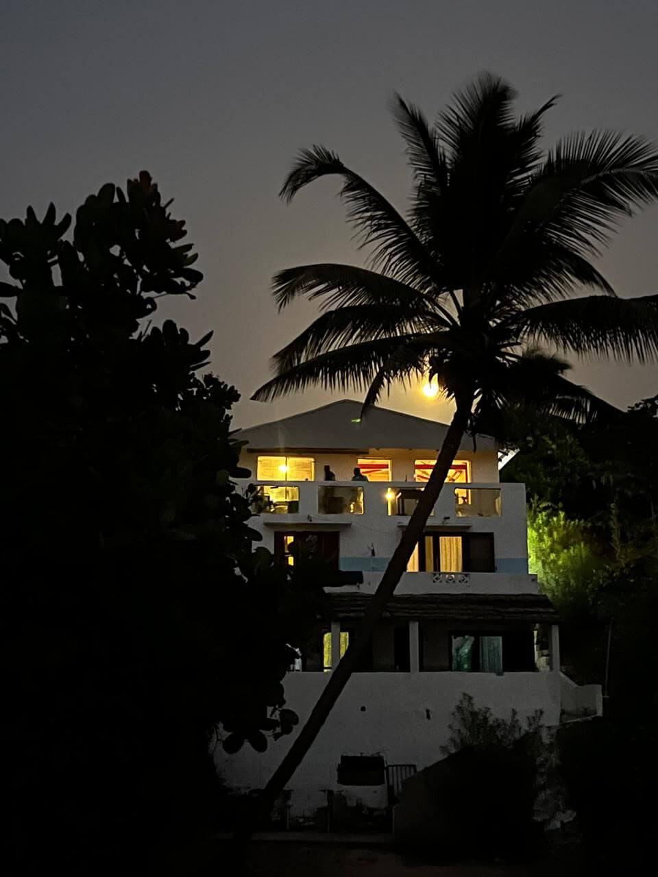 Façade éclairée du Campement Buhanor au crépuscule ou de nuit, avec des palmiers en silhouette, offrant une ambiance chaleureuse et tropicale à Cap-Skirring