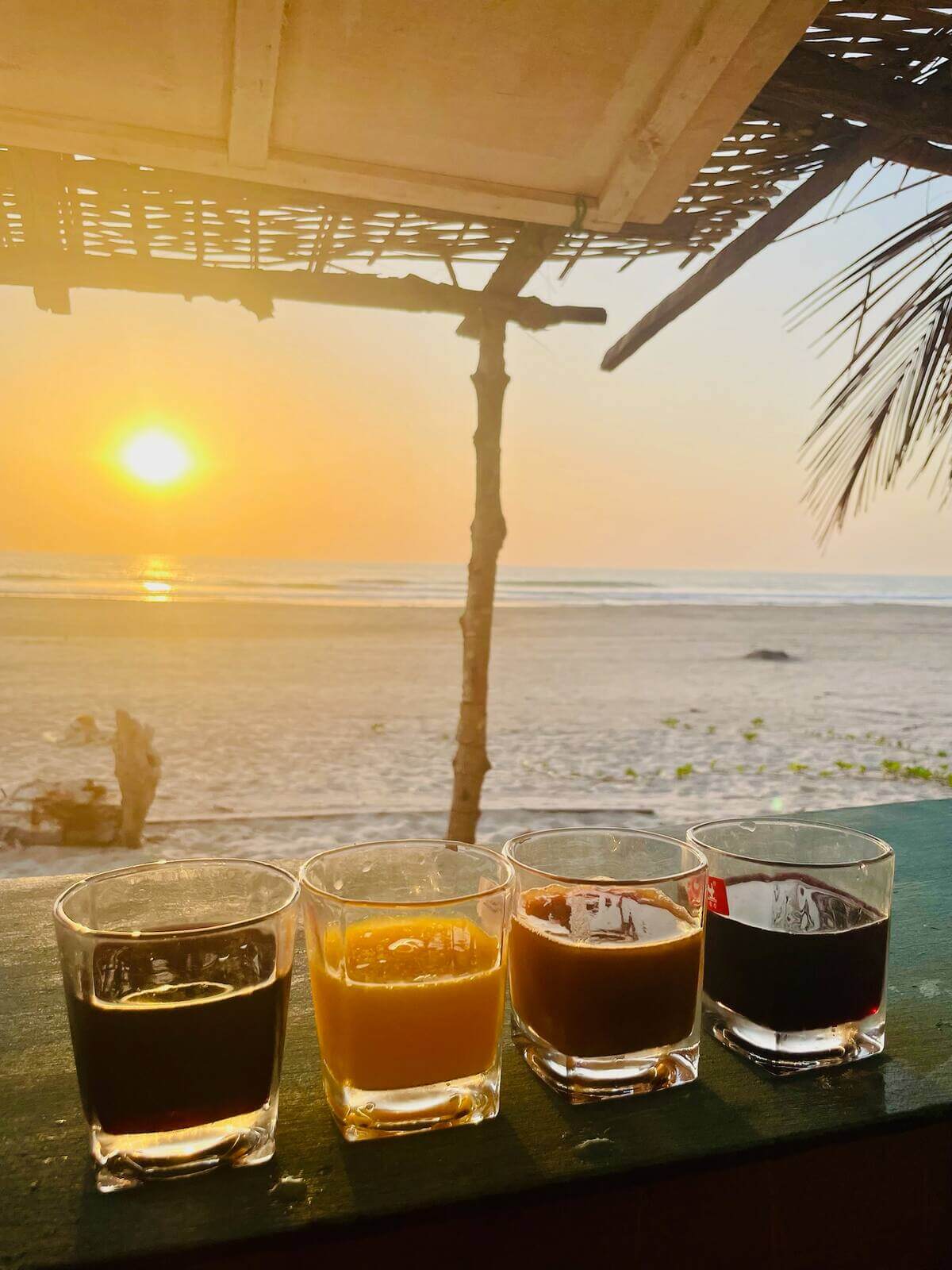 Quatre verres de jus locaux et boissons fraîches sur une table, avec le coucher de soleil et l'océan de Cap-Skirring en arrière-plan, au Campement Buhanor