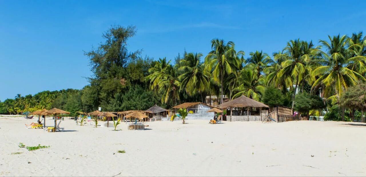 Vaste plage de sable blanc à Cap-Skirring bordée de palmiers et de quelques structures ombragées, un cadre paradisiaque typique de la Casamance près du Campement Buhanor.